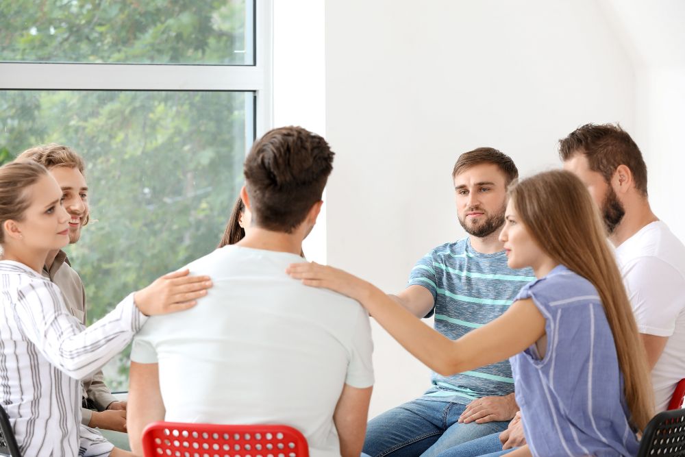 Sad man looks downward as others in his group therapy session comfort